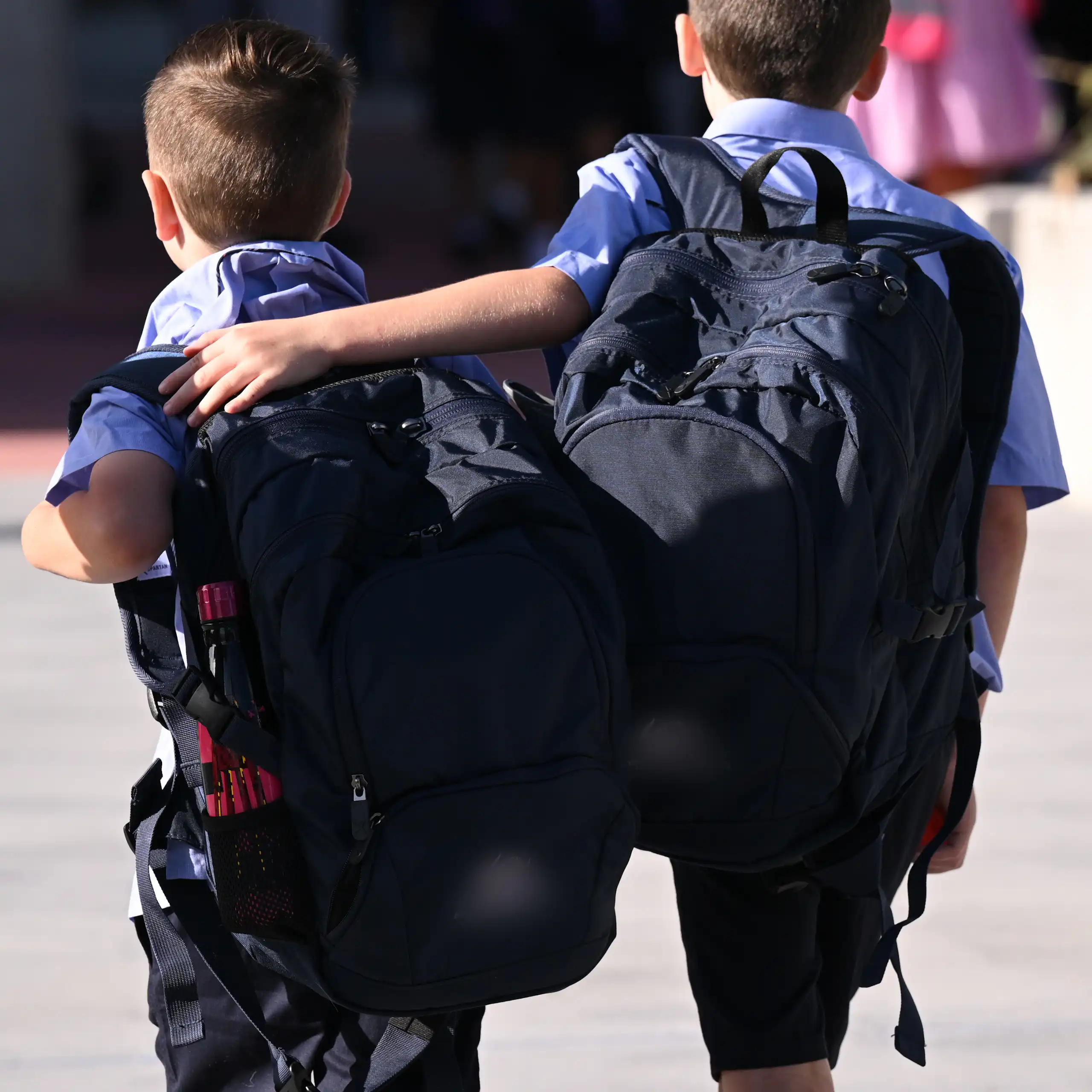 Two young students with backpacks walk along. One has an arm around the other.