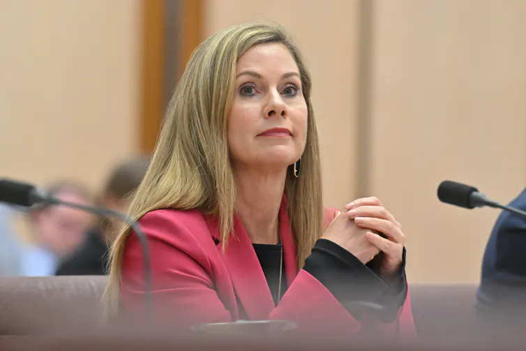 Woman with blond hair wearing a pink blazer, sitting at a table with her hands crossed.
