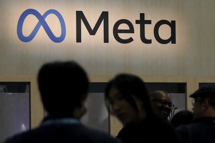 Two people standing in front of the Meta logo which features a blue figure eight on its side.