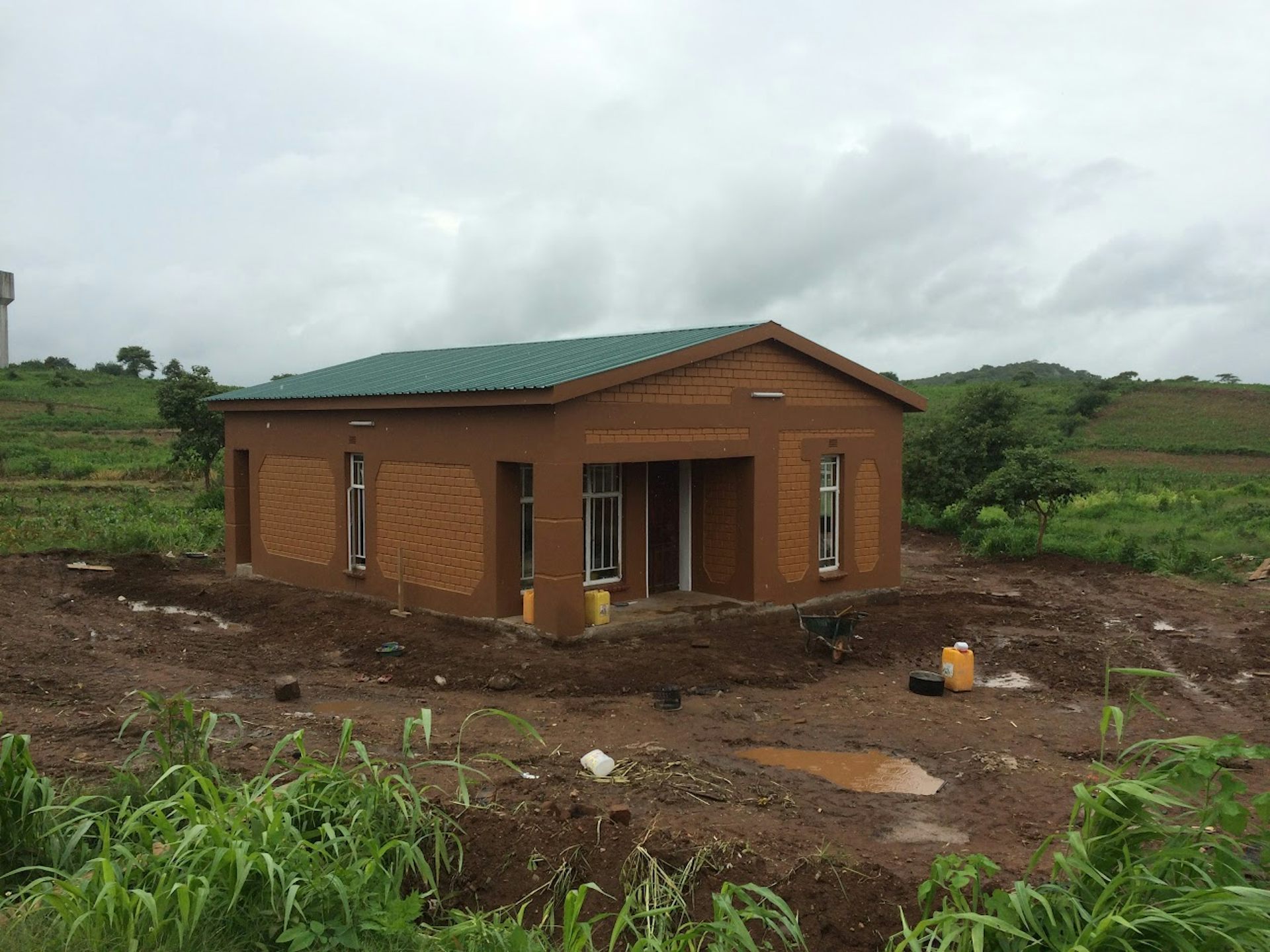 A brown house with a green roof and a verandah built on an open plot of land with hills in the background