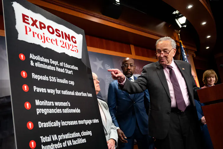 An older man wearing a grey suit points his finger at a large poster that says 'Exposing Project 2025'