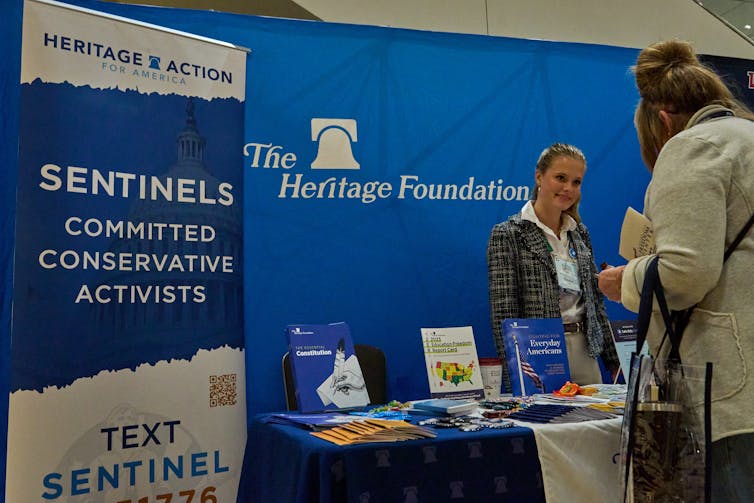 A white woman wears a blazer and stands at a table in front of a blue backdrop that says 'The Heritage Foundation'