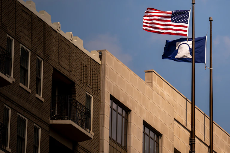Two flags - one American and the other blue with a white bell - fly next to a large stone building.