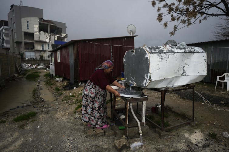 A woman washes a metal dish under an outdoor water tank next to a red metallic structure. Damaged concrete buildings are seen in the background