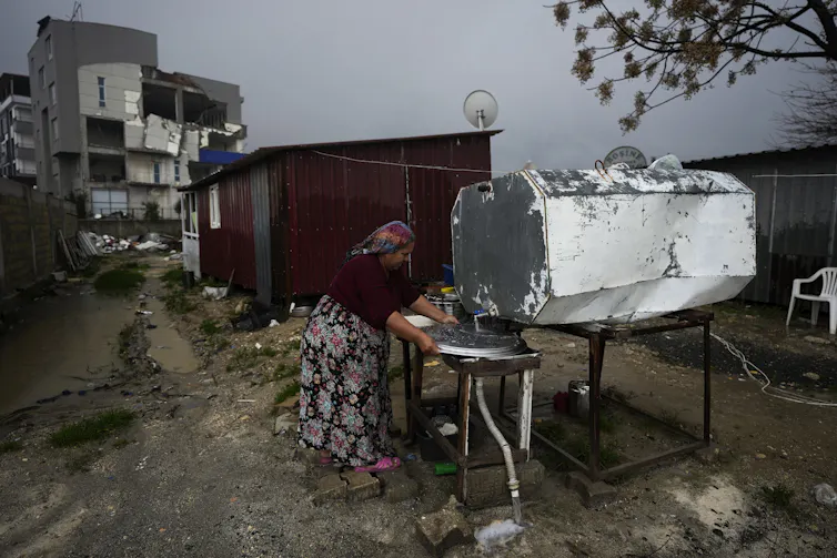 A woman washes a metal dish under an outdoor water tank next to a red metallic structure. Damaged concrete buildings are seen in the background