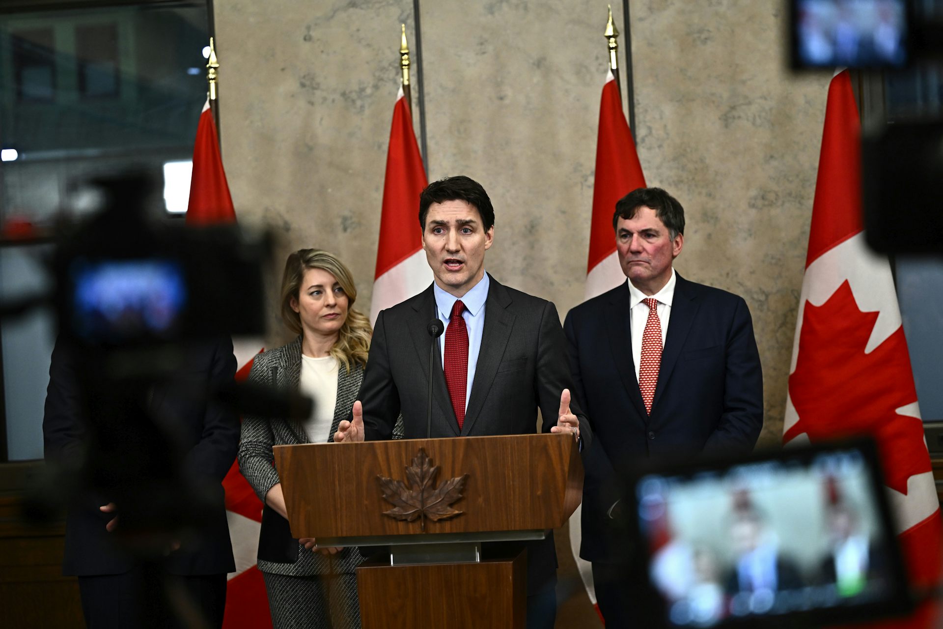 Un homme blanc en costume s'exprime depuis un podium, avec des drapeaux canadiens derrière lui. Il est entouré d'une femme blanche et d'un homme blanc.