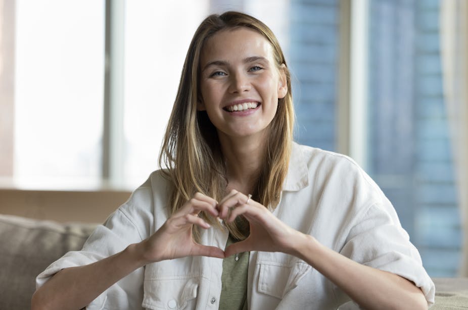 A smiling young woman looks at the camera, forming a heart shape with her hands.