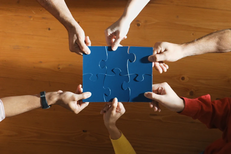 Overhead view of six people's hands holding a large blue puzzle together over a conference table