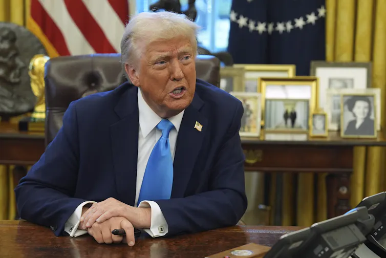 A man with orange-tinged skin and white hair speaks to someone off camera while sitting at a desk
