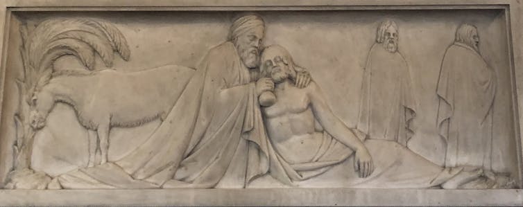 A carving in stone shows a man in robes kneeling beside a half-naked man on the ground as two other figures walk by.