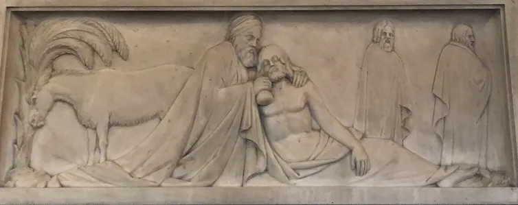 A carving in stone shows a man in robes kneeling beside a half-naked man on the ground as two other figures walk by.