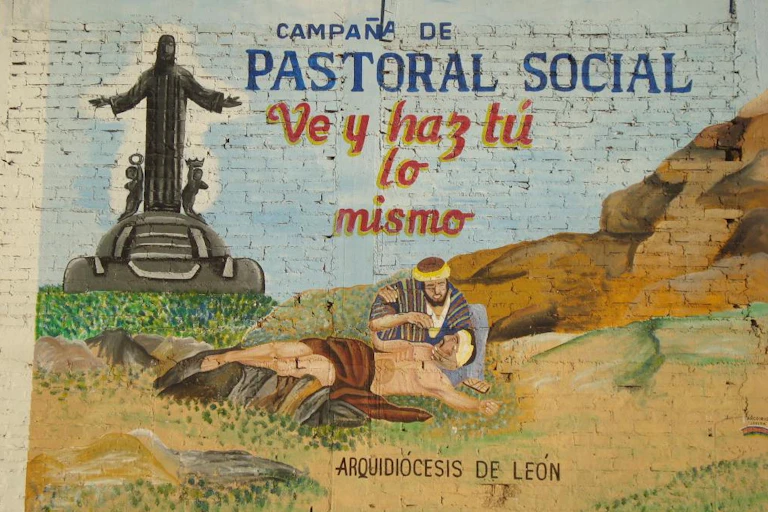 A mural and words painted on whitewashed bricks depicts a man kneeling as he offers water to a bandaged man on the ground.