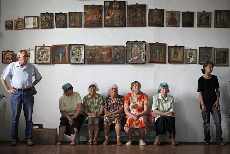 Several women sit on a bench against a white wall that displays many religious paintings.
