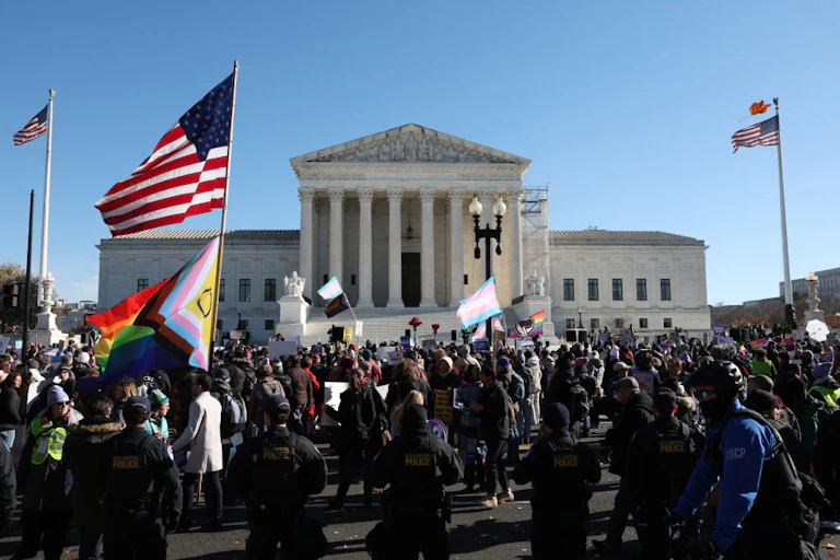 Large group of people gathered before the U.S. Supreme Court, some holding rainbow and trans flags; a line of police in tactical gear stand in the foreground