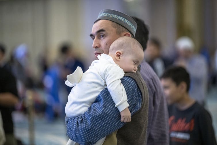 A man in a blue shirt and dark vest holds a baby in a white outfit in the middle of a large room full of people.