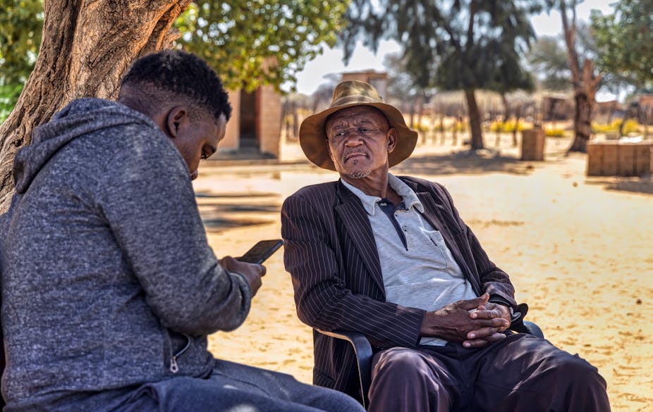 A young person looks at a cellphone while an older man looks on with a slightly suspicious expression
