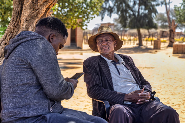 A young person looks at a cellphone while an older man looks on with a slightly suspicious expression