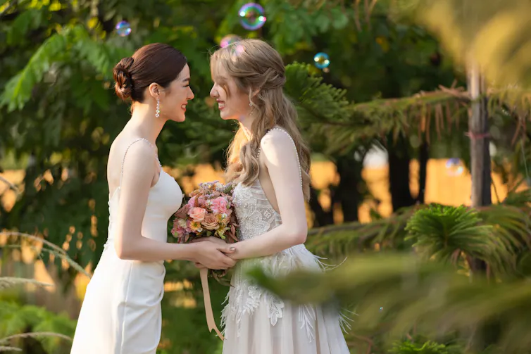 Two women in wedding dresses stand facing each other, holding hands and smiling joyfully. One holds a bouquet of pink and peach flowers. Sunlight filters through the trees, with bubbles floating in the air.