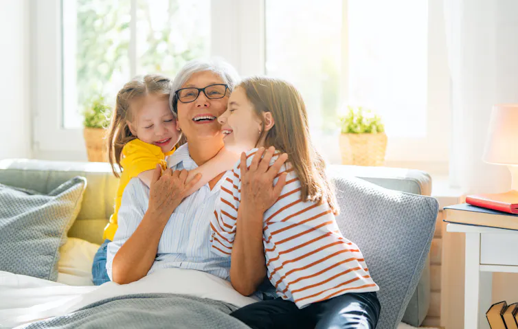 une femme aux cheveux gris assise sur un sofa et encadrée par deux petites filles
