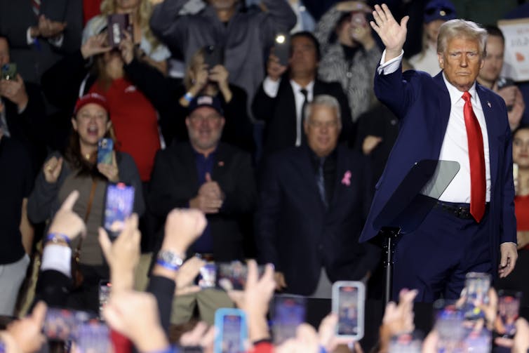 A man wearing a blue suit and white shirt waves to a crowd of people smiling and holding up phones.