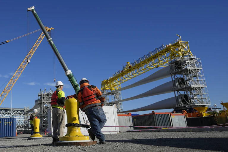 Two workers stand on a dock with three large wind turbine blades on a ship in front of them.