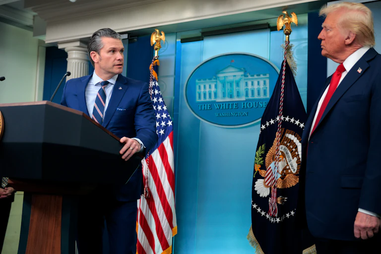 Two men in blue suits, one standing at a lectern, in front of two flags.
