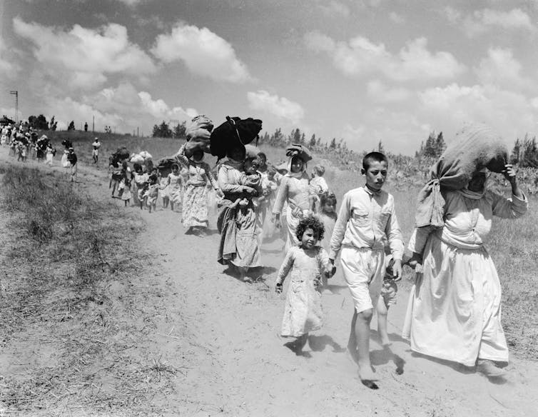 A line of children and women walk along a dusty track, many with bundles on their heads.