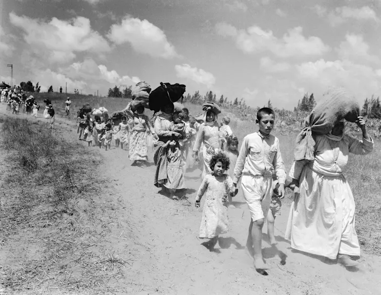 A line of children and women walk along a dusty track, many with bundles on their heads.