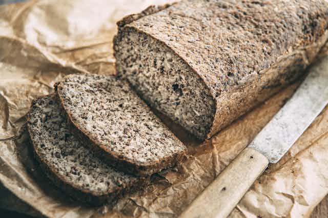A loaf of brown bread with a bread knife nearby is displayed on brown wrapping paper.