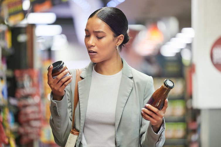 a woman in a supermarket aisle holding two bottles of sauce and reading the label of one