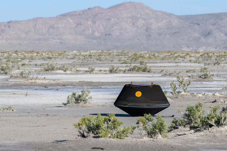 Black capsule sitting in the sun in the middle of a desert.