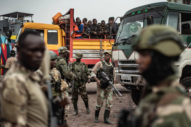 Men in army fatigues and holding guns surround trucks filled with men, some dressed in army fatigues and others in civilian clothing