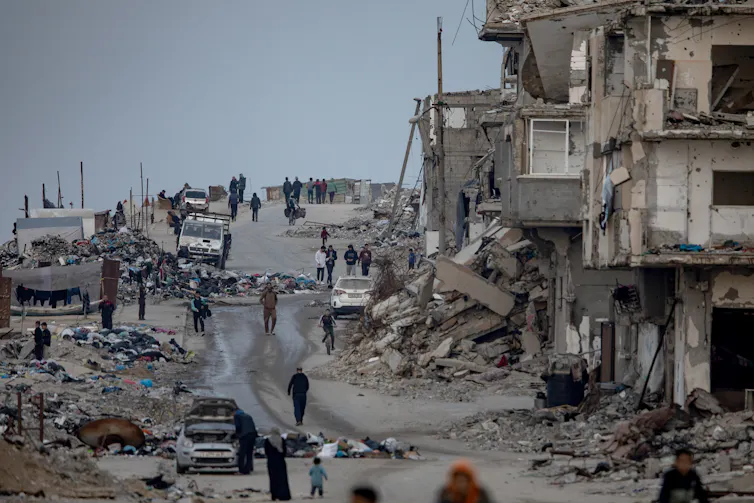 Palestinians walk through a street among the rubble of destroyed buildings.