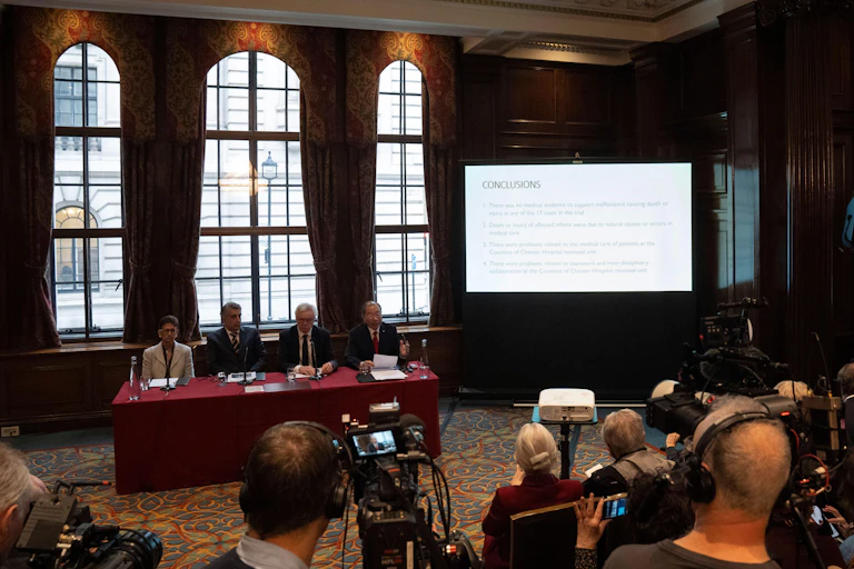Press conference in room with tall windows behind and a screen showing conclusions reached by the expert panel.