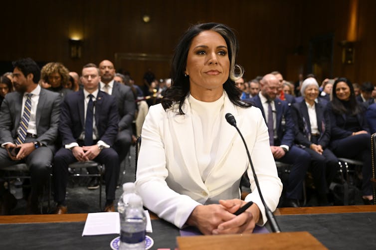 A woman in a white suit sits at a table, with an audience behind her.