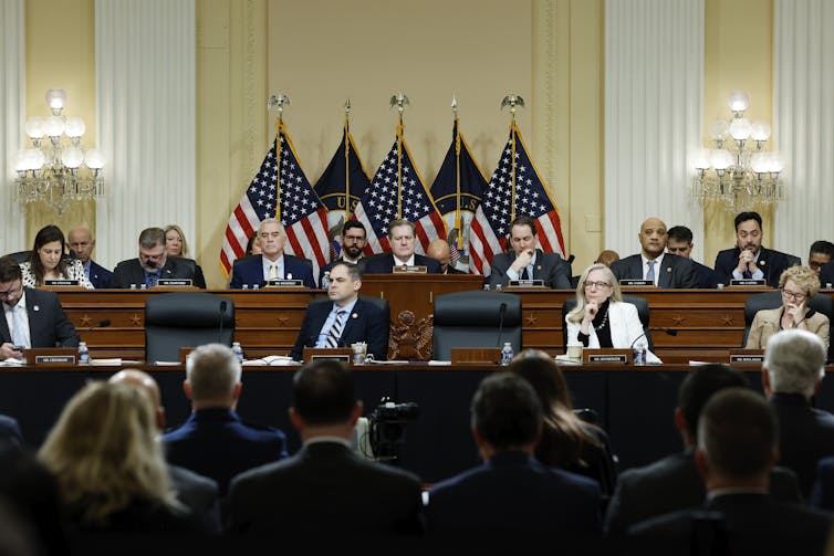 A group of people in business attire sit at a wooden dais backed by U.S. flags.