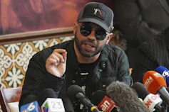 A bearded man with sunglasses and wearing a ball cap speaks at a news conference.