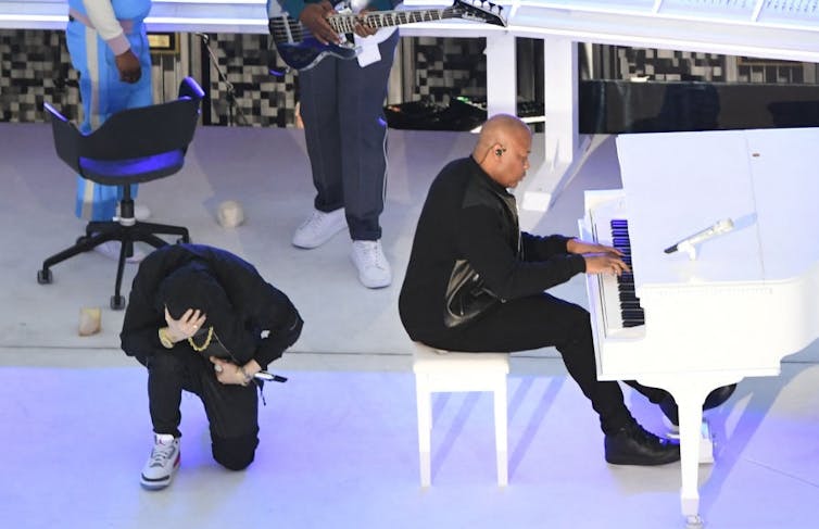 Rapper dressed in black takes a knee next to a bald man sitting on a white piano bench and playing a white piano.