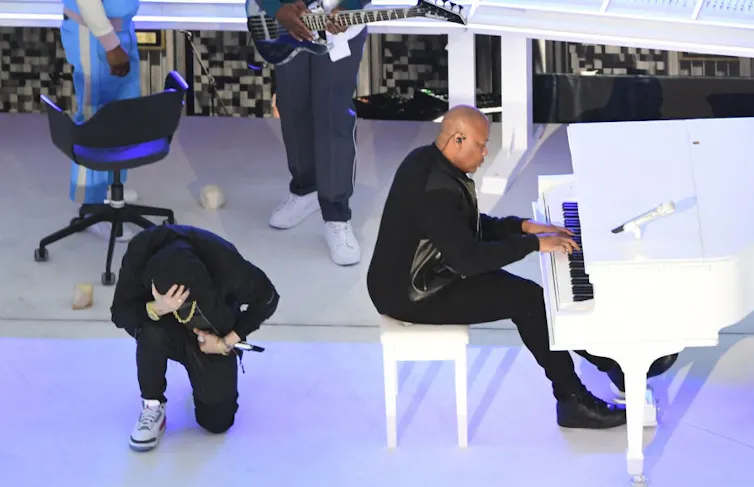 Rapper dressed in black takes a knee next to a bald man sitting on a white piano bench and playing a white piano.