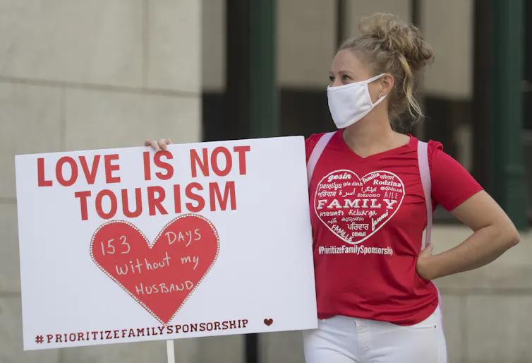 A woman in a face mask wearing a red shirt carries a sign reading: love is not tourism 153 days without my husband