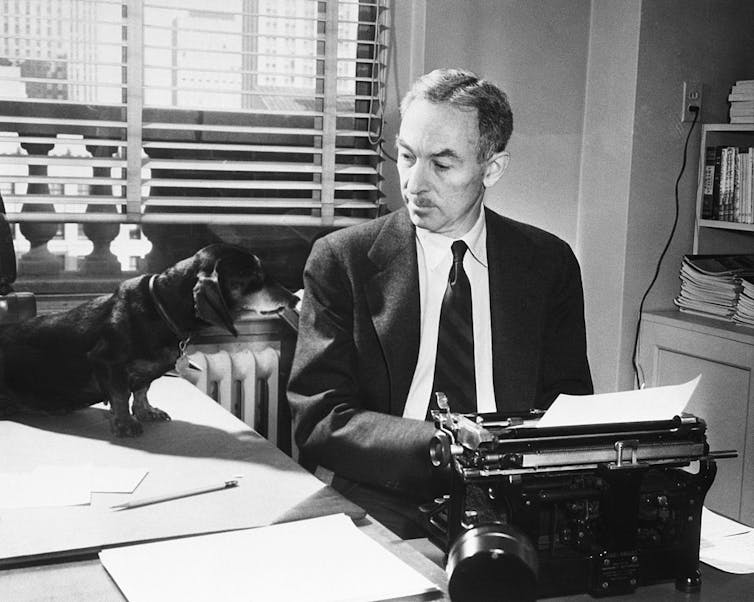 Middle-aged man wearing suit and tie seated at typewriter looking at a small dog perched on a table to his right.