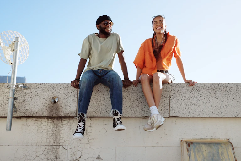 A young man and woman sit on a ledge on a rooftop together smiling.