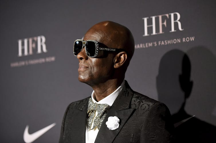 A black man in a pair of sunglasses, suit and a bowtie stands in front of a wall that has the Harlem Fashion Row collective and Nike logos on it