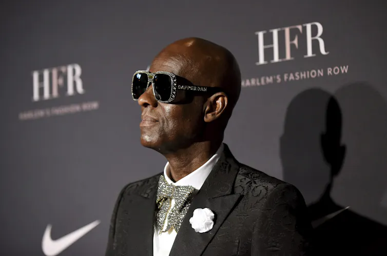 A black man in a pair of sunglasses, suit and a bowtie stands in front of a wall that has the Harlem Fashion Row collective and Nike logos on it