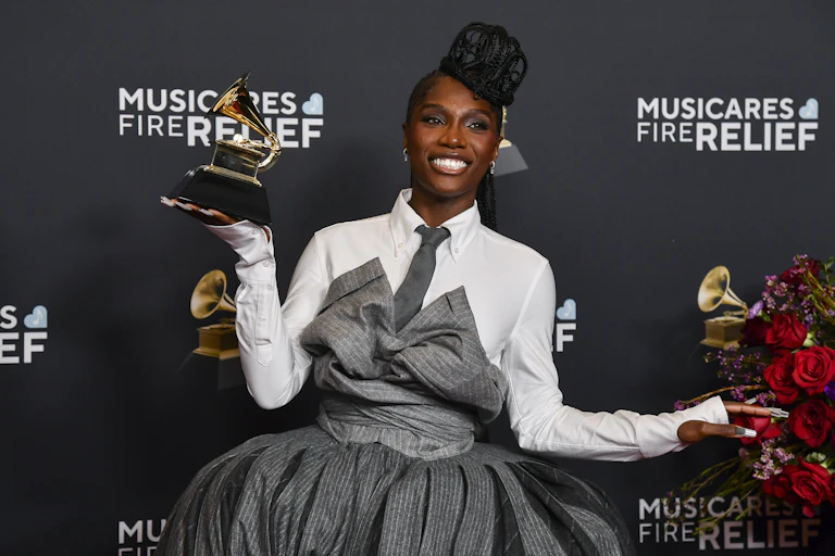 A black woman with her hair braided into a crown shape in a grey and white suit dress holds a grammy award in one hand while smiling