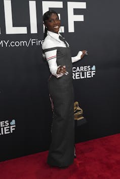 A black woman with long, elaborately braided hair wears a grey off-the-shoulder corset suit dress with exaggerated hips paired with a white dress shirt and grey tie