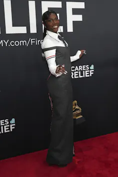 A black woman with long, elaborately braided hair wears a grey off-the-shoulder corset suit dress with exaggerated hips paired with a white dress shirt and grey tie