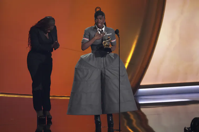 A black woman with her hair braided into loops wears a cropped, short-sleeved grey jacket with a tie, paired with dramatically structured balloon pants