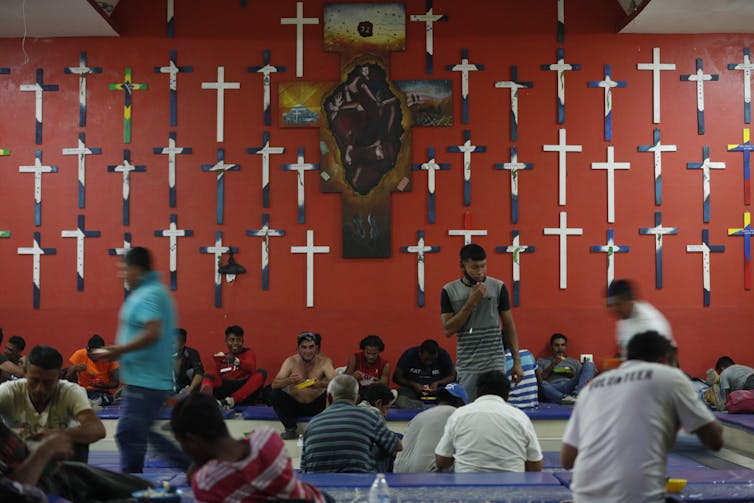 Whether or not Christians must prioritize take care of migrants up to for fellow voters has been debated for hundreds of years 1 People are eating while seated in rows on a carpeted floor. Mounted on the red wall behind them are several crucifixes.