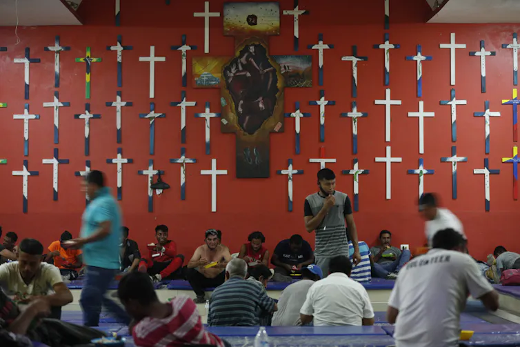 People are eating while seated in rows on a carpeted floor. Mounted on the red wall behind them are several crucifixes.